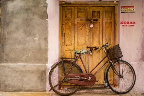 A black roadster leans against a golden wooden door, the tailor's number painted on the pink wall beside it.