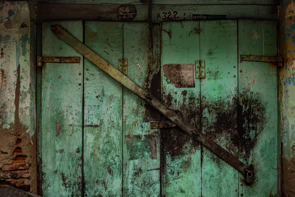 A teal wooden door crossed by a heavy diagonal brace, rust bleeding through its paint.
