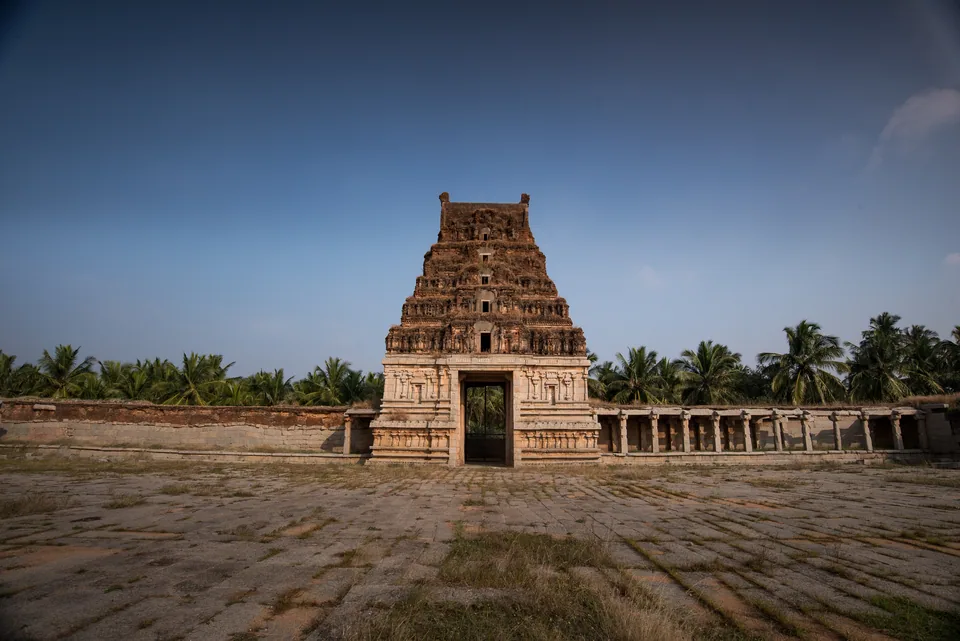 A temple gateway and its long pillared corridor across a courtyard of worn stone, palms beyond.