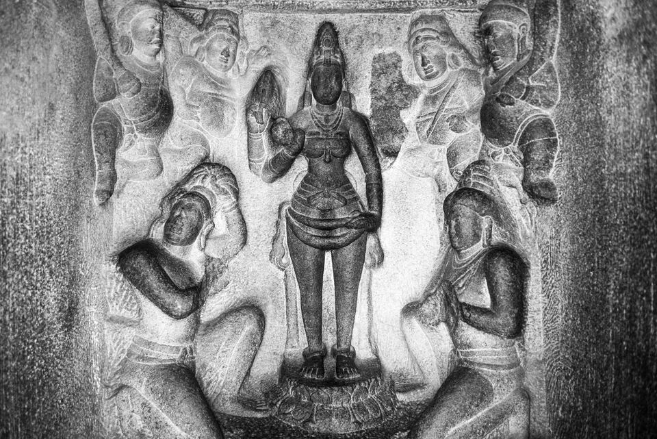 A rock-cut goddess stands among attendant figures in a carved panel, rendered in silvery monochrome.