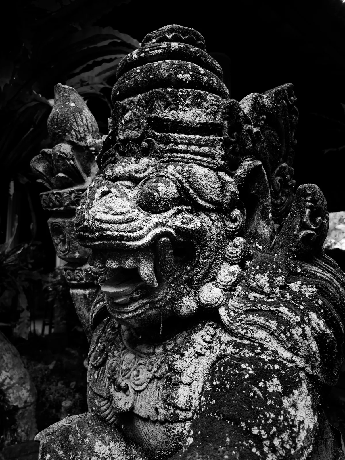 A moss-dark guardian in carved stone, its fierce face softened by age.