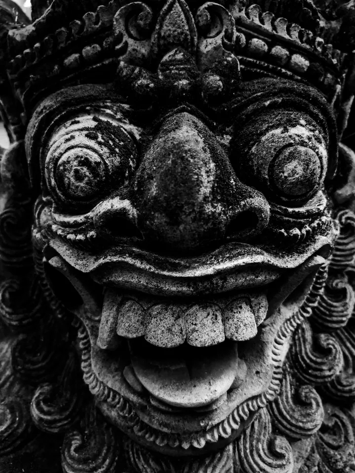 A demon mask carved in stone, all bulging eyes and wild grin: the protective face of a Balinese temple in deep monochrome.