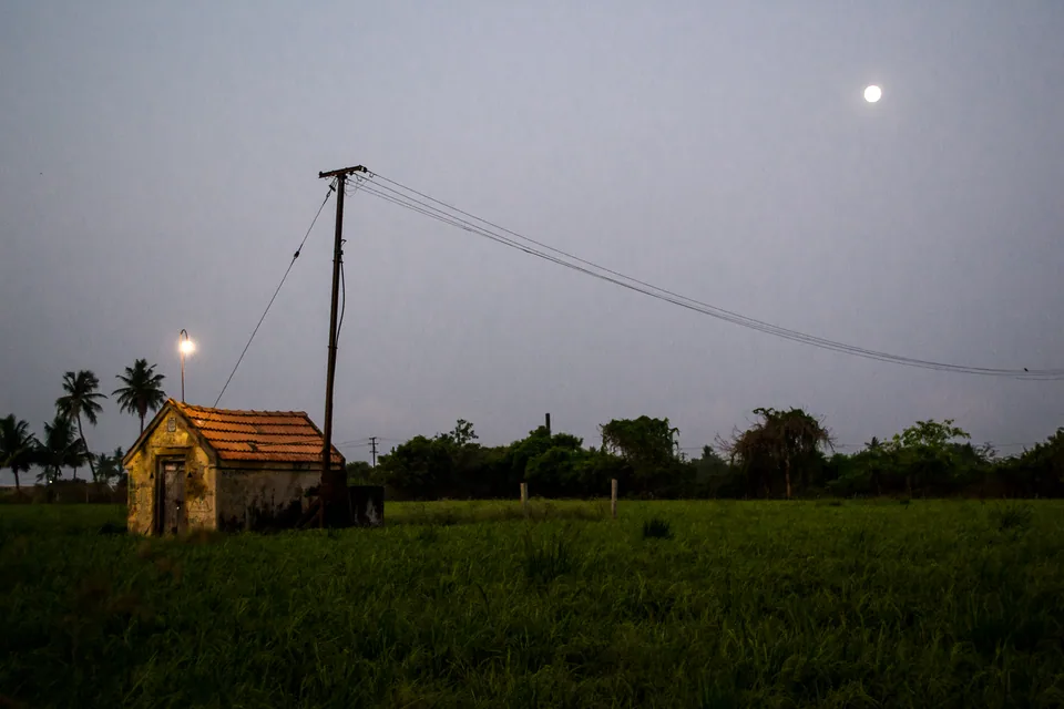 A small tiled house, an electric pole and a rising moon over green fields at dusk.