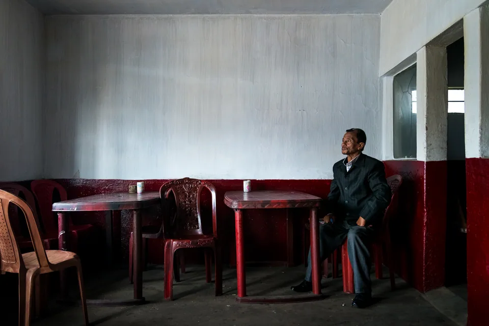 A man in a dark coat waits in an empty tea room of red chairs and pale walls.