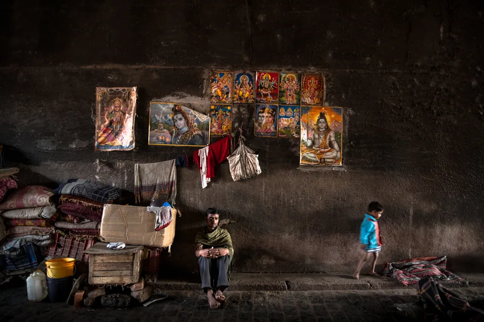 A family's corner beneath a bridge, bright posters of gods on the wall above.