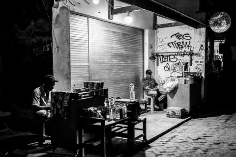 A small shop at night, its keeper in the doorway and graffiti on the shutter.